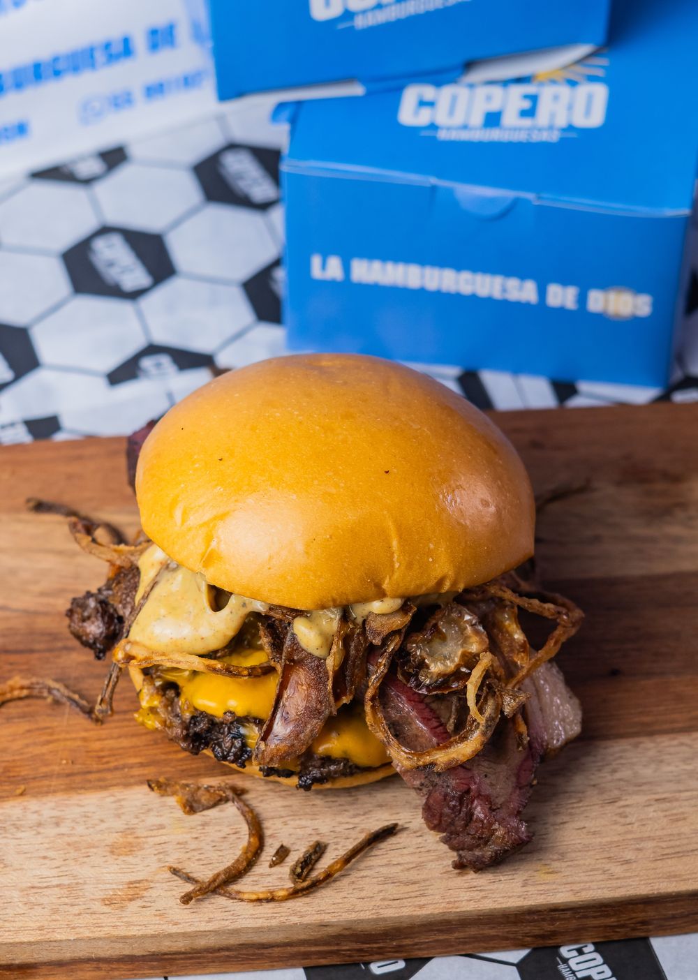 Hambúrguer da Copero Hamburguesas sobre tábua, com as embalagens azuis ao fundo