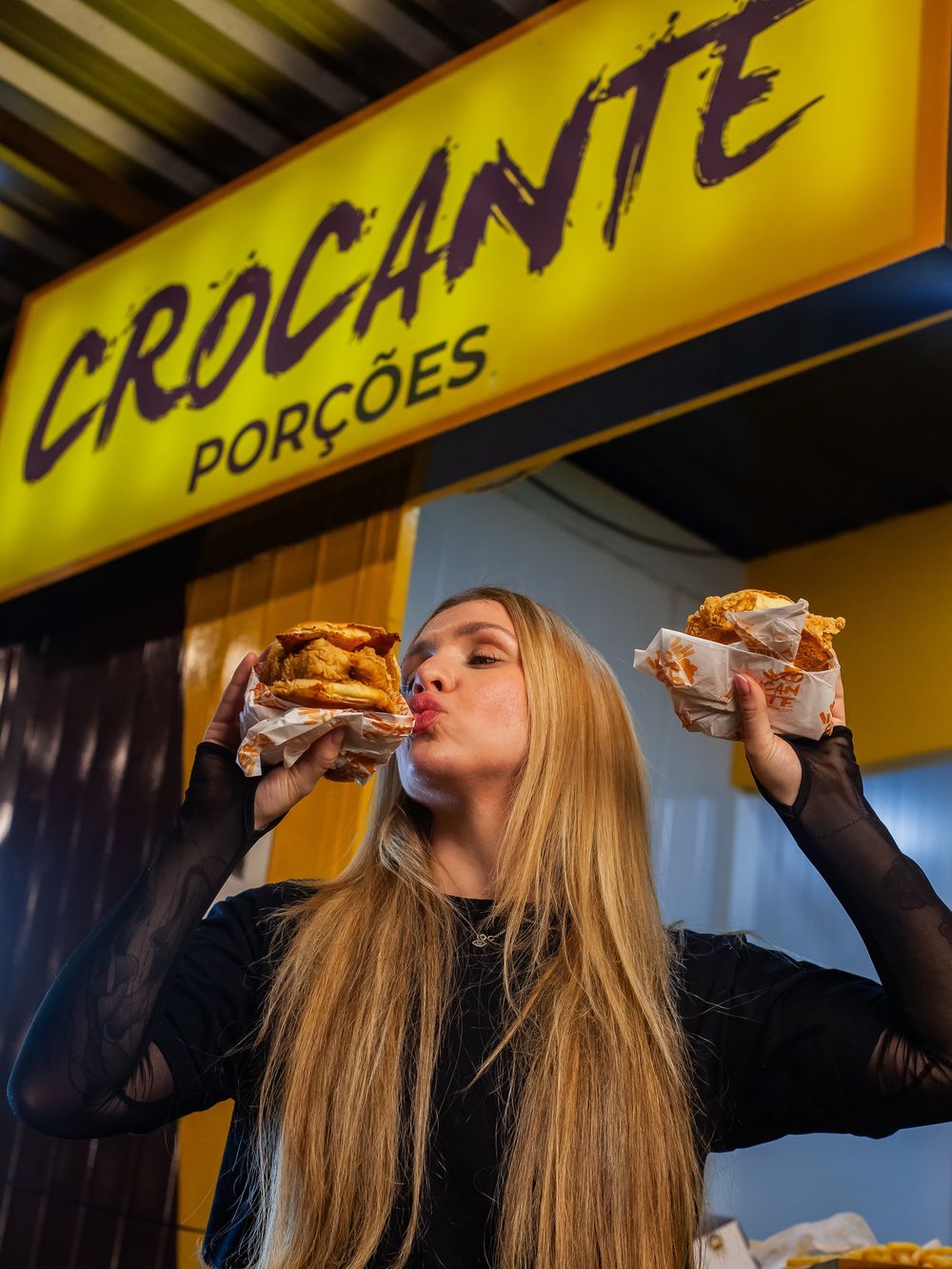 Modelo segurando dois lanches em frente ao letreiro da Crocante Porções