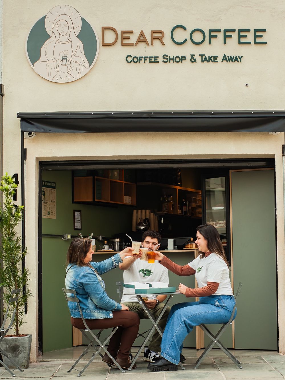 Três pessoas brindando com bebidas na fachada do Dear Coffee